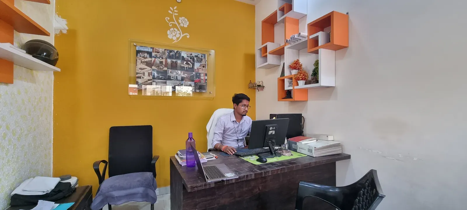 The Miraj Construction office on Ring Road: mustard wall, orange display shelves and a framed collage of project photos